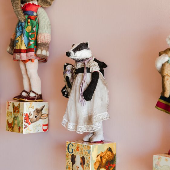 A collection of vintage, hand-stitched badger and dog figures stands on colourful alphabet blocks against a pink wall.