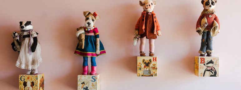 A collection of small, handcrafted animal figurines – a badger, fox, pig, and bear – are mounted on a pale pink wall. Each figure stands on a coloured alphabet block (G, S, H, B).