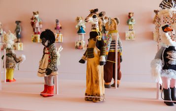 A gallery display featuring a collection of diverse, handcrafted animal figures. The giraffe stands prominently, surrounded by a monkey and goose, all in various vintage garments.