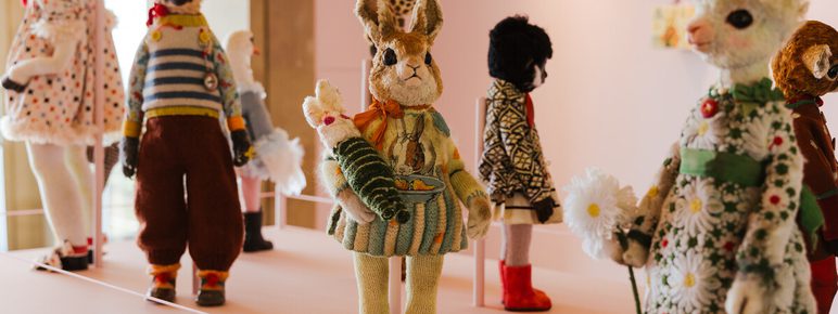 A display of handcrafted, plush animal dolls is presented on a pale pink wall. The collection includes a rabbit, bear, llama, and zebra, each with unique outfits and accessories.
