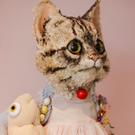 A close-up shot of a vintage, plush cat toy dressed in a miniature pink dress and adorned with a red button and beaded necklace. A small, stuffed mouse rests beside it.