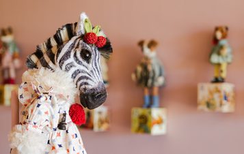 A close-up shot of a stuffed zebra wearing a frilled collar and a patterned dress, with red berries adorning its neck. Several decorative figurines are visible in the background.