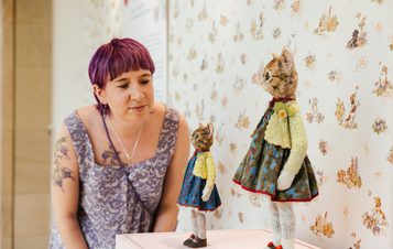 A woman examines two miniature knitted cat figures displayed on a plinth, set against a floral wallpaper.