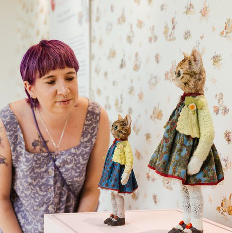 A woman examines two miniature knitted cat figures displayed on a plinth, set against a floral wallpaper.
