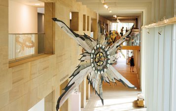 A large, weathered metal sculpture resembling a star hangs in a modern museum corridor. People are visible in the background.