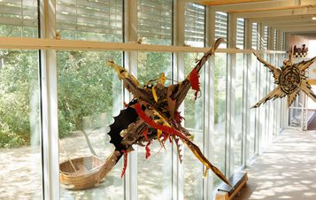 A colourful, intricate mask hangs on a wall within a modern building, alongside a small wooden boat. Large windows offer a view of a leafy garden outside.