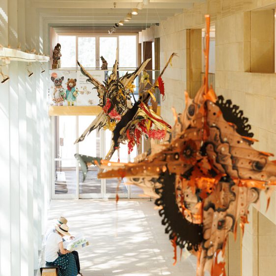 A person sits sketching in a bright, airy corridor of a museum. Large, colourful, sculptural artworks hang from the ceiling, creating a dynamic display.