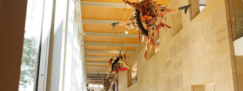 A contemporary art installation featuring a large, multi-layered sculpture hangs within a light-filled corridor of a building. The ceiling is clad in wood panelling.