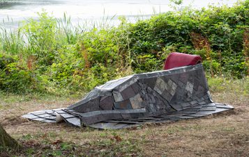 A stone sculpture of a seated figure, with a red cushion, situated by a lake with reeds and foliage in the background.