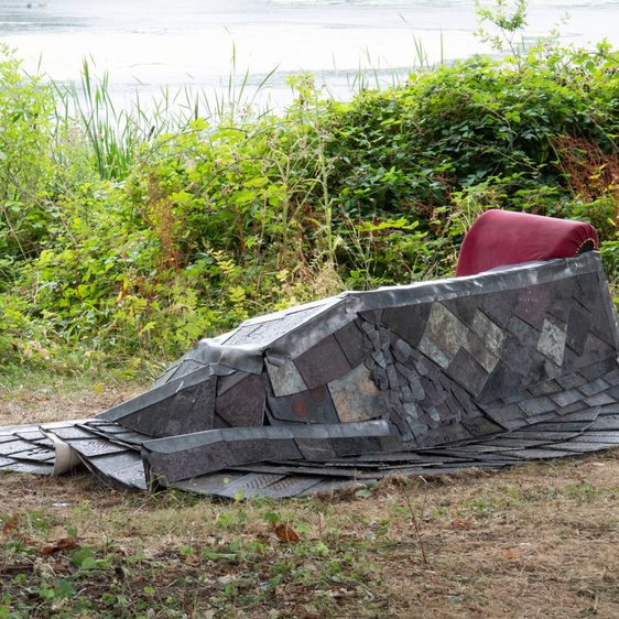 A stone sculpture of a seated figure, with a red cushion, situated by a lake with reeds and foliage in the background.