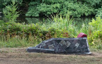 A modern, geometric sculpture resembling a boat rests on a grassy bank beside a river, with a red cushion visible. Lush greenery forms the backdrop.