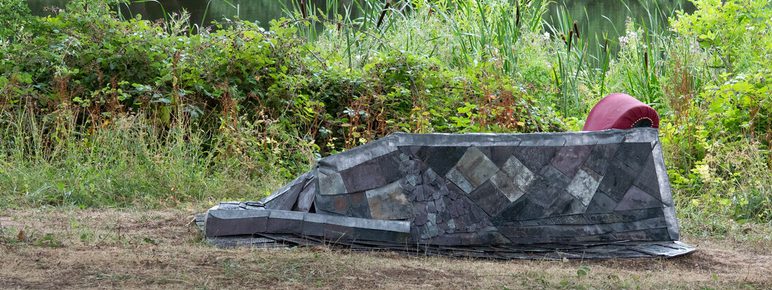A modern, geometric sculpture resembling a boat rests on a grassy bank beside a river, with a red cushion visible. Lush greenery forms the backdrop.