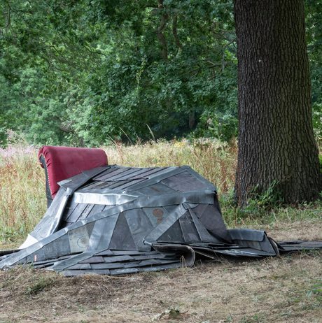 A metal sculpture resembling a seated figure rests on a grassy bank beside a large tree in a woodland setting. A red armchair is positioned beside the sculpture.