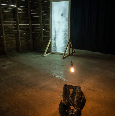 An art installation in a dimly lit space features a large, framed abstract artwork on a wooden easel. Below, a sculptural piece made of dark, textured material rests on the floor. A single light bulb hangs overhead, casting soft shadows.