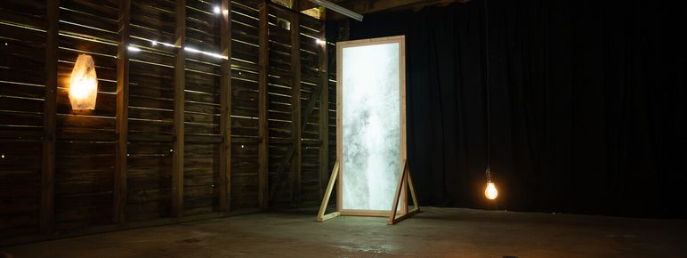 An empty, rustic room with wooden walls and a bare floor. A large, framed mirror stands upright, reflecting light. A single light bulb hangs from the ceiling, and a dark, abstract sculpture lies on the floor, adding an artistic touch to the space.