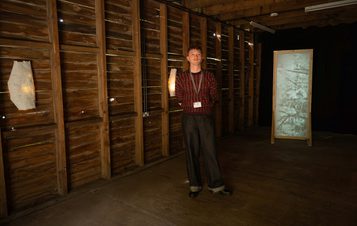 A person stands in a dimly lit room with wooden walls, wearing a patterned sweater and dark pants. They hold a piece of paper and look towards the camera. A lighted panel with a translucent surface is positioned nearby, casting a soft glow.