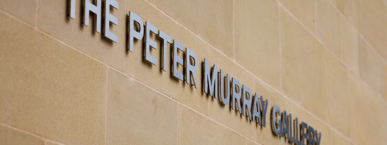 A close-up view of a wall featuring the sign for "The Peter Murray Gallery." The letters are metallic and mounted on a light-colored stone surface, creating a modern and elegant appearance.