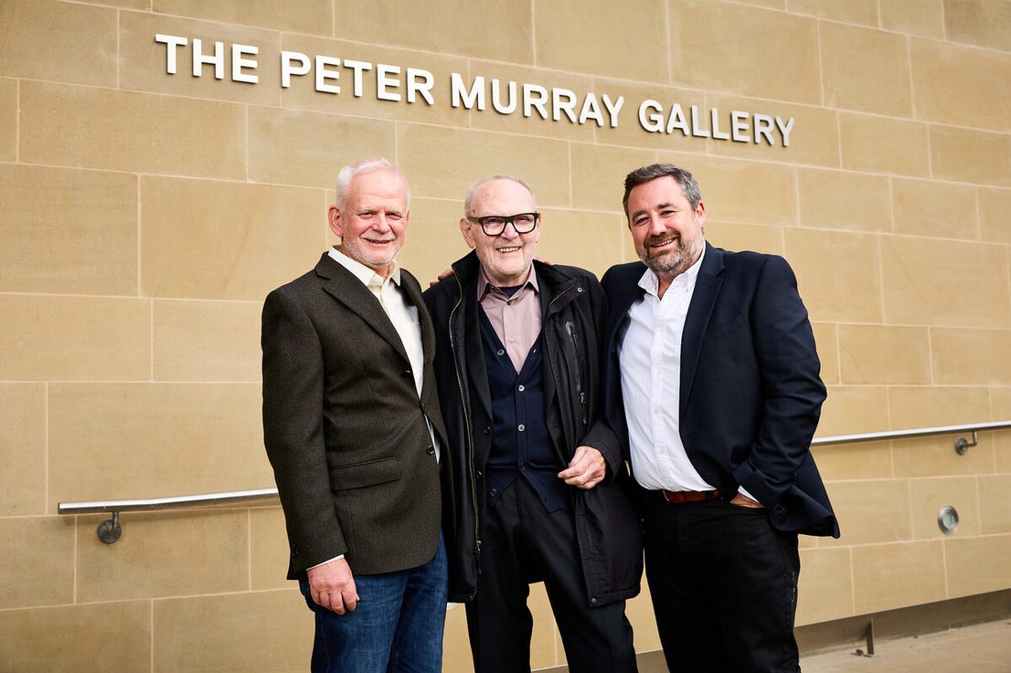 Three men stand in front of a stone wall with the text "THE PETER MURRAY GALLERY" displayed above. They are smiling and dressed in smart casual attire, with one man wearing glasses. The setting appears to be an outdoor gallery entrance.