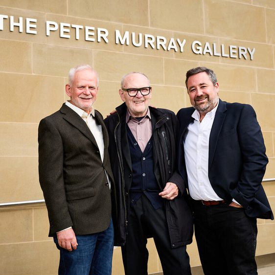 Three men stand in front of a stone wall with the text "THE PETER MURRAY GALLERY" displayed above. They are smiling and dressed in smart casual attire, with one man wearing glasses. The setting appears to be an outdoor gallery entrance.
