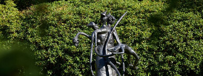 A striking bronze sculpture of a figure entwined with animals, set against a lush green backdrop. The figure appears dynamic and expressive, showcasing intricate details and a harmonious connection with nature. Sunlight casts gentle shadows around it.