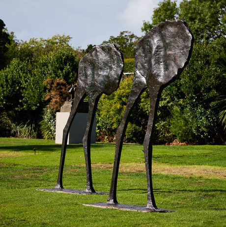 Two tall, abstract sculptures resembling elongated leaves or figures stand on a grassy area, surrounded by lush greenery. The sculptures are made of a dark material, contrasting with the vibrant landscape and cloudy sky in the background.