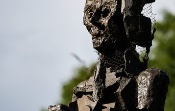 A weathered metal sculpture of a seated figure, featuring a textured surface and a skeletal appearance. The figure has a mesh-like head and is positioned against a backdrop of greenery and a blue sky, conveying a sense of contemplation.
