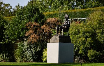 A bronze statue of a seated figure, deep in thought, is positioned on a square pedestal amidst lush greenery. The surrounding plants include various shrubs and trees, creating a serene garden atmosphere.