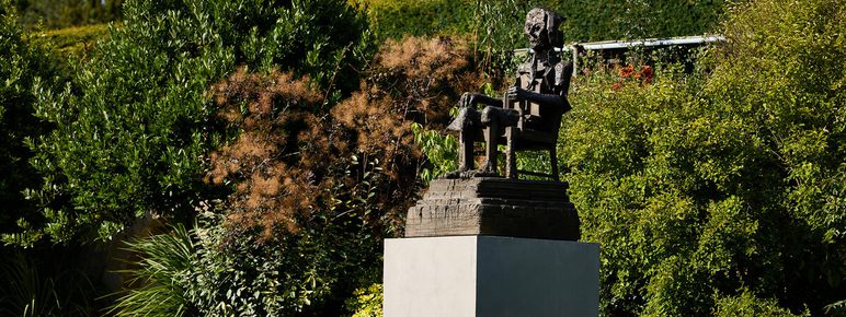 A bronze statue of a seated figure, deep in thought, is positioned on a square pedestal amidst lush greenery. The surrounding plants include various shrubs and trees, creating a serene garden atmosphere.