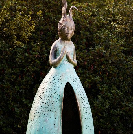 A large, whimsical sculpture of a figure with rabbit-like ears, standing with hands clasped. The figure has a textured, turquoise dress with perforations, set against a backdrop of lush greenery. The artwork blends fantasy and nature.