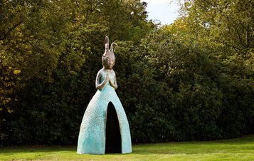 A large, whimsical sculpture of a figure with elongated features and a dress-like form stands in a green park. The figure has a serene expression and is surrounded by lush trees, creating a tranquil outdoor setting.