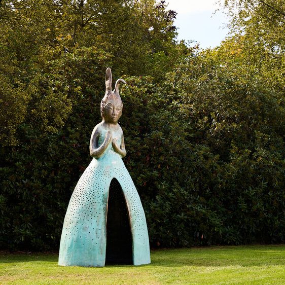 A large, whimsical sculpture of a figure with elongated features and a dress-like form stands in a green park. The figure has a serene expression and is surrounded by lush trees, creating a tranquil outdoor setting.