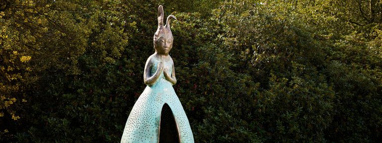 A large, whimsical sculpture of a figure with elongated features and a dress-like form stands in a green park. The figure has a serene expression and is surrounded by lush trees, creating a tranquil outdoor setting.