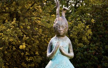 A bronze sculpture of a girl with elongated, horn-like hair stands in front of lush greenery. She has a serene expression and is posed with her hands clasped together, wearing a dress with perforated patterns. The background features vibrant yellow foliage.