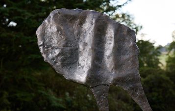 A close-up of a textured bronze sculpture resembling a leaf or abstract form, with visible grooves and a natural patina. The background features blurred greenery, suggesting an outdoor setting. The sculpture stands prominently against the landscape.