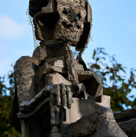 A weathered sculpture of a seated figure with a skeletal face, made from various materials including wood and wire mesh. The figure has a textured surface and is positioned against a blue sky with greenery in the background.