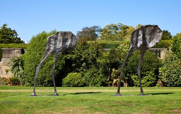 Two abstract bronze sculptures resembling elongated figures with flat, irregular heads and thin legs stand on a grassy area. They are set against a backdrop of lush greenery and trees under a clear blue sky.