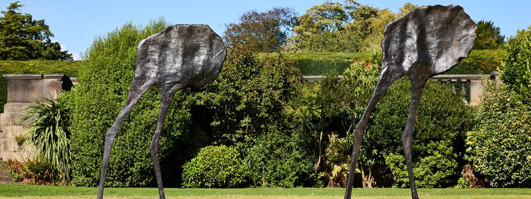 Two abstract bronze sculptures resembling elongated figures with flat, irregular heads and thin legs stand on a grassy area. They are set against a backdrop of lush greenery and trees under a clear blue sky.