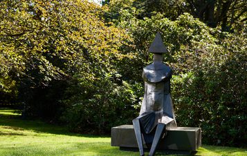 A large, abstract sculpture of a seated figure with a triangular head and geometric body sits on a stone bench. It is surrounded by lush greenery and dappled sunlight, creating a serene outdoor setting.