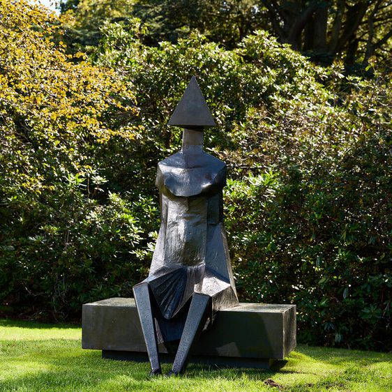 A large, abstract sculpture of a seated figure with a triangular head and geometric body sits on a stone bench. It is surrounded by lush greenery and dappled sunlight, creating a serene outdoor setting.