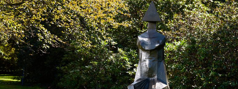 A large, abstract sculpture of a seated figure with a triangular head and geometric body sits on a stone bench. It is surrounded by lush greenery and dappled sunlight, creating a serene outdoor setting.