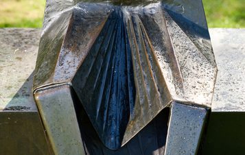 A close-up of a metallic sculpture featuring sharp, angular folds and a glossy surface. Water droplets cling to the textured metal, reflecting light. The background is blurred, emphasizing the intricate details of the sculpture.