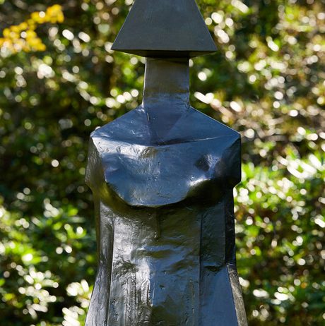 A close-up of a modern sculpture featuring a stylized figure with a triangular head and a smooth, abstract body. The sculpture is set against a blurred background of greenery, highlighting its dark, metallic finish.
