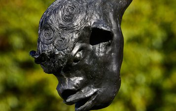 A close-up of a dark, abstract sculpture featuring a stylized human head with exaggerated features. The head is elongated and appears to be emerging from a curved form, set against a blurred green background of foliage.