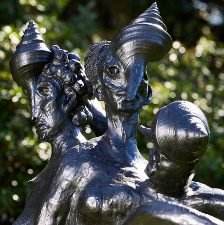 A bronze sculpture featuring three stylized figures with elongated heads and spiral horn-like features. The figures are intertwined, showcasing intricate details in their expressions and hair, set against a blurred green background.