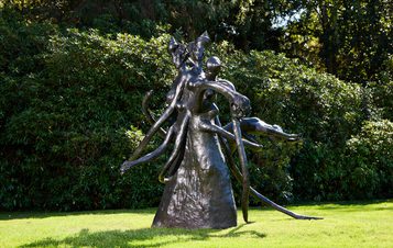 A large bronze sculpture depicting abstract human figures intertwined, with elongated limbs and flowing forms. The artwork is set against a backdrop of lush greenery, highlighting its dynamic shapes and textures in natural light.