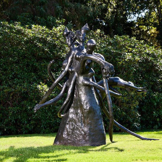 A large bronze sculpture depicting abstract human figures intertwined, with elongated limbs and flowing forms. The artwork is set against a backdrop of lush greenery, highlighting its dynamic shapes and textures in natural light.