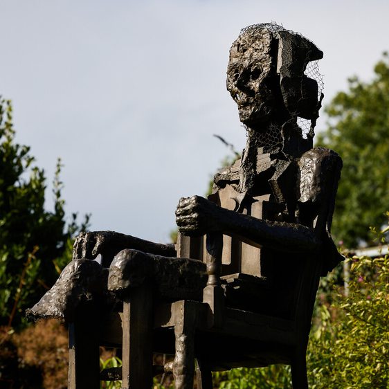 A weathered metal sculpture of a seated figure with a fragmented head and body, surrounded by greenery. The figure appears contemplative, with a textured surface reflecting light, set against a cloudy sky.
