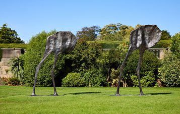 Two abstract bronze sculptures resembling elongated figures with flat, leaf-like heads stand on a grassy area. They are positioned apart, surrounded by lush greenery and trees under a clear blue sky.