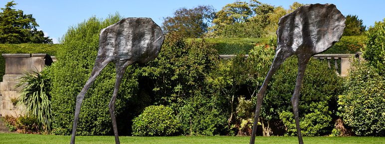 Two abstract bronze sculptures resembling elongated figures with flat, leaf-like heads stand on a grassy area. They are positioned apart, surrounded by lush greenery and trees under a clear blue sky.