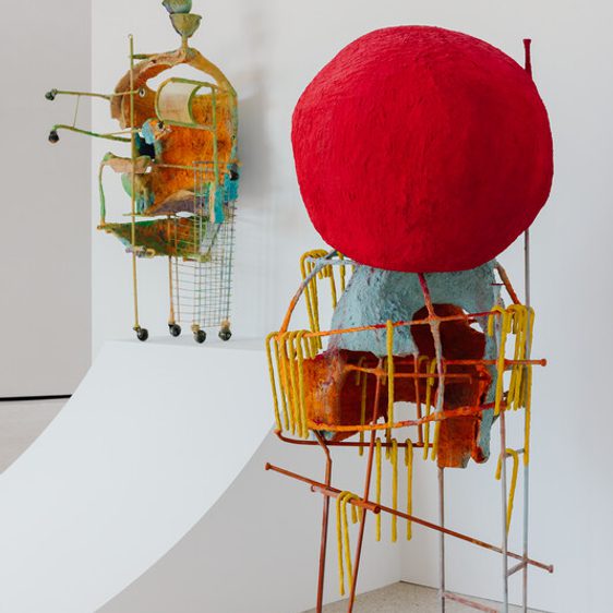 Two abstract sculptures are displayed in a gallery. One features a large red spherical head atop a colorful, cart-like base, while the other resembles a whimsical machine with various shapes and textures, both set against a white wall.